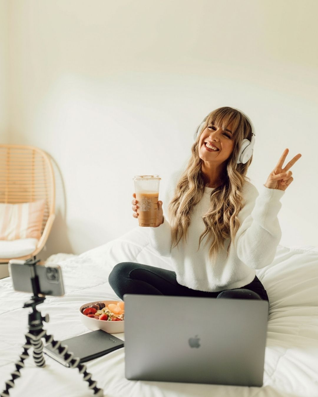 Sammy sitting on a bead with a glass in her hand and a laptop on the bed.She is wearing a white sweatshirt and has one white headphones